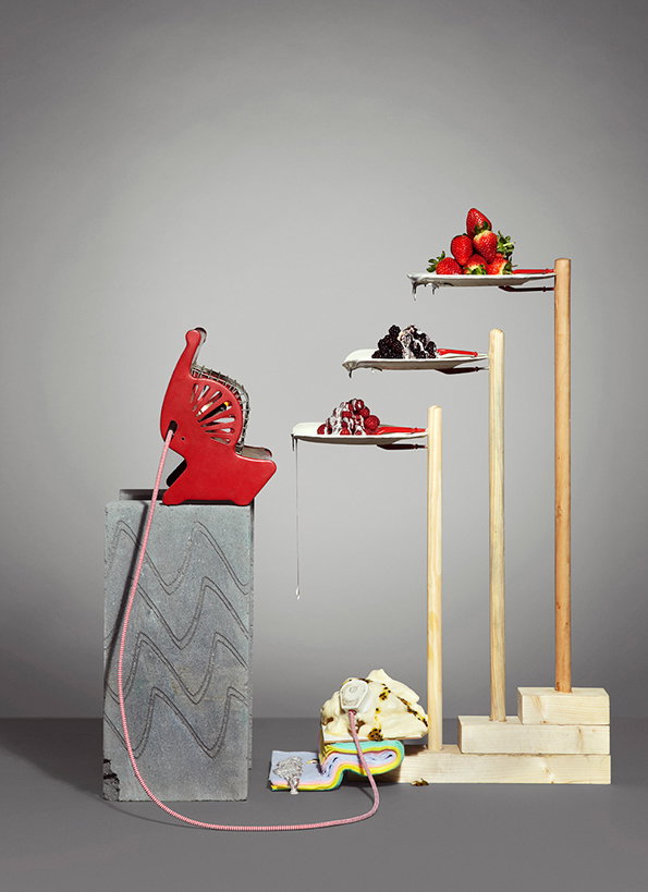 A red, stylised telephone sits atop a grey concrete block. A pink cord leads down to a small, colourful sculpture on the floor. To the right, three white plates are held at different heights by wooden poles. The plates hold strawberries, blackberries and a red dessert.