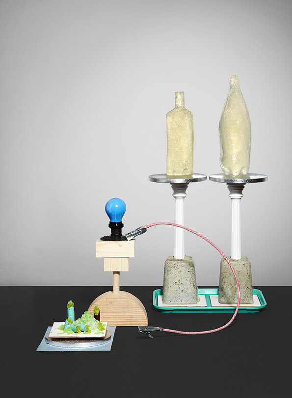 A sculpture featuring two translucent, yellowish bottles resting on white pedestals with concrete bases. A pink cable connects the pedestals to a wooden structure topped with a blue lightbulb and a small, colourful landscape model.