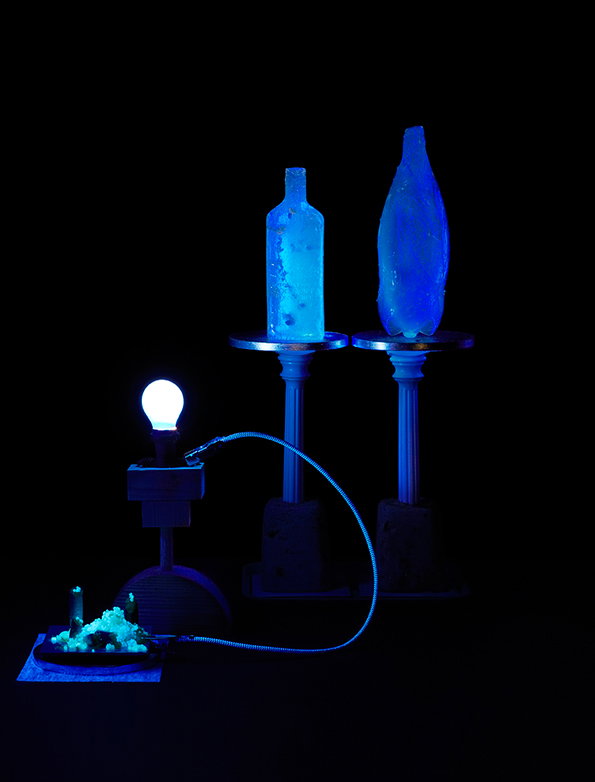 Two blue, translucent sculptures resembling bottles stand on tall pedestals against a black background. A glowing lightbulb and a small, crystalline structure are connected by a thin, luminous blue cable.