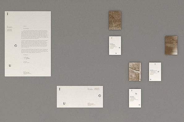 A collection of white papers and brown textured cards are arranged across a grey wall. The papers feature minimalist layouts with the letters “I”, “U” and “O” placed around the edges of the pages.