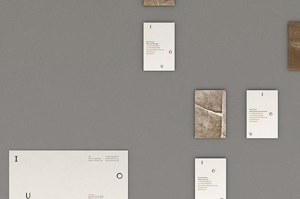 Several white business cards and textured brown stone tiles are arranged in a loose grid on a grey wall. The cards feature minimalist typography with the letters “I”, “O” and “U” spaced vertically.