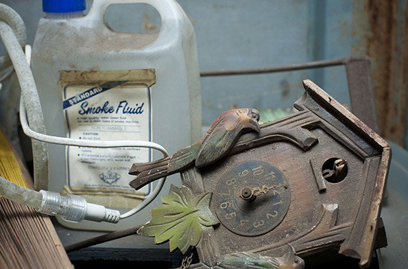 A dusty, ornate clock with a bird ornament lies on its side. Behind it sits a large plastic jug with a label that reads “Standard Smoke Fluid”.
