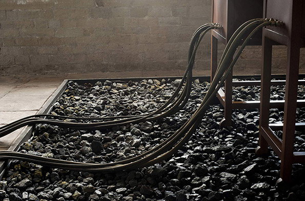 Thick black cables snake across a bed of dark coal. They lead from the floor into the base of a dark wooden cabinet.