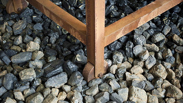 A close-up of a rusted metal frame resting on a bed of grey and brown jagged stones. The frame forms a V-shape, with a small wheel visible at the base of one leg.