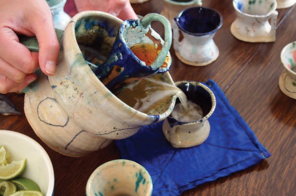 A person pours a pale liquid from a large, glazed ceramic pitcher into a small matching cup. The pitcher and cups feature blue and earthy tones and sit on a wooden table.