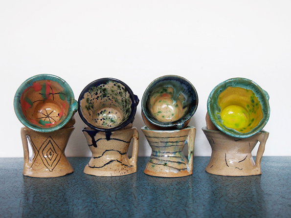 Four handmade ceramic cups sit in a row on a blue textured surface. Each cup has a unique glaze and a chunky, sculpted base with a small handle.