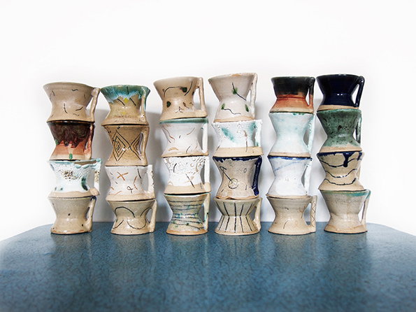 Six ceramic sculptures stand in a row on a blue surface against a white background. Each piece is a vertical stack of four or five vessel-like forms in various neutral and blue glazes.
