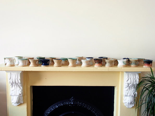 A row of handmade ceramic cups in various earthy glazes and shapes lines a pale yellow mantelpiece. Below them, a black ornate fireplace is framed by white decorative carvings.