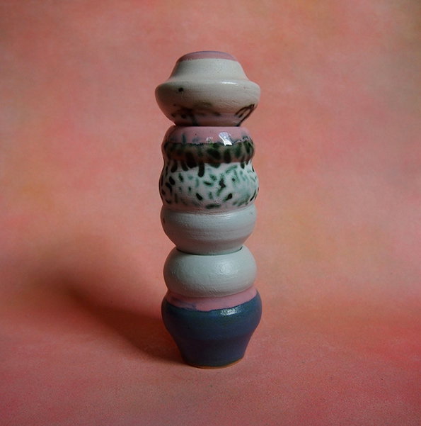 A ceramic sculpture made of stacked, rounded shapes in shades of pink, white and blue. The piece stands against a soft, blurred pink background.