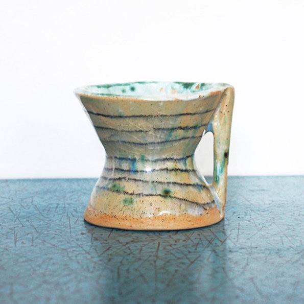 A ceramic coffee dripper with a handle, glazed in earthy tones of beige and blue. It sits on a textured teal surface against a plain white background.