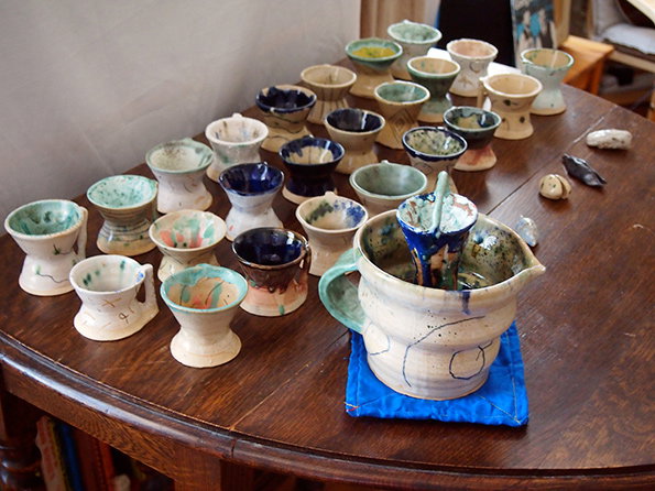 A collection of handmade ceramic cups and bowls in various sizes and glazes are arranged across a dark wooden table. A larger pitcher sits in the foreground on a blue fabric square, holding a few smaller vessels.