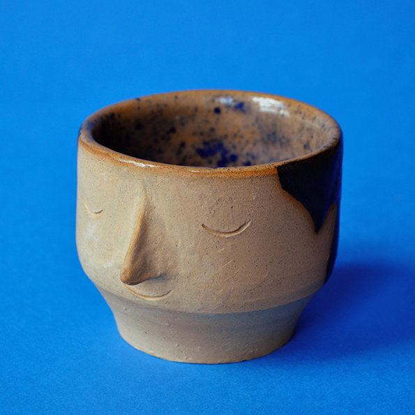 A ceramic cup shaped like a face, with a prominent nose and closed eyes. The piece is glazed in earthy brown tones with a splash of dark blue on one side, set against a bright blue background.