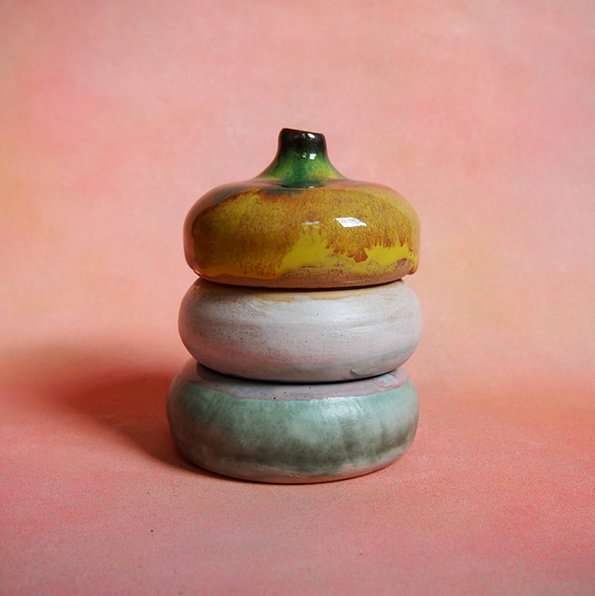 A ceramic sculpture of three stacked, rounded forms against a soft pink background. The top piece is yellow and green, resembling a small fruit, resting on two pale, glazed bowls.