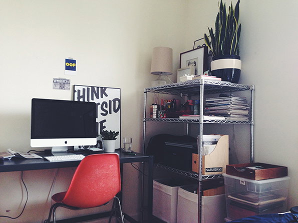 A home office setup with a computer on a dark desk and a bright red chair. Beside the desk, a metal shelving unit holds a snake plant, a lamp, and various organisers.