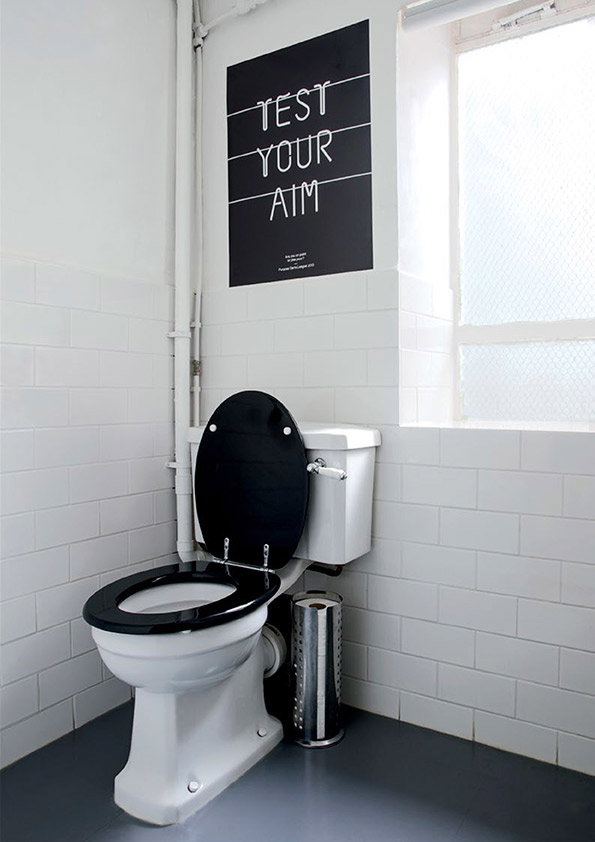 A white toilet with a black seat and lid stands against a white tiled wall. Above it, a black poster with white lines reads “TEST YOUR AIM”.