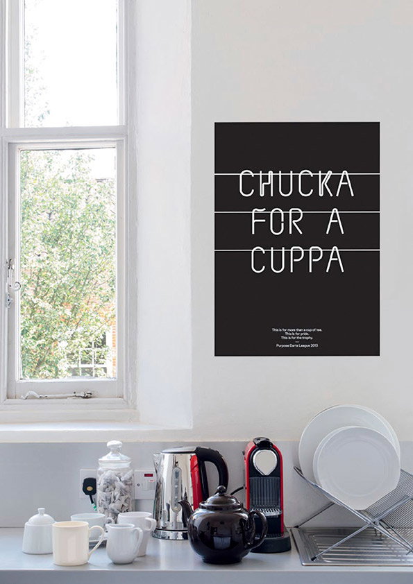 A black poster on a white wall reads “CHUCKA FOR A CUPPA” in white capital letters. Smaller text at the bottom mentions the “Purpose Drive League 2013”. Below the poster, a kitchen counter holds a kettle, a black teapot and a stack of white plates.