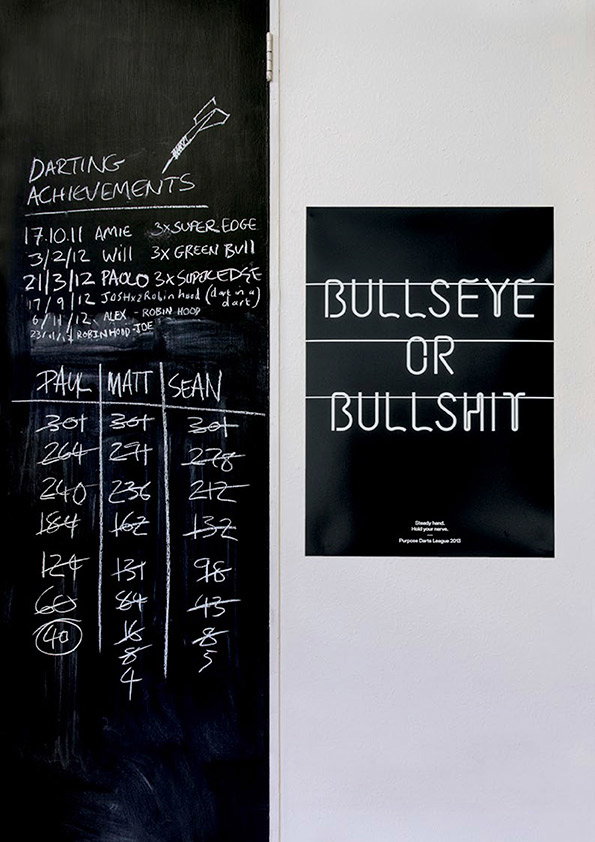 A chalkboard lists “Darting Achievements” and a table of scores for Paul, Matt and Sean. Beside it, a black poster reads “Bullseye or Bullshit” in a thin, white sans-serif typeface.
