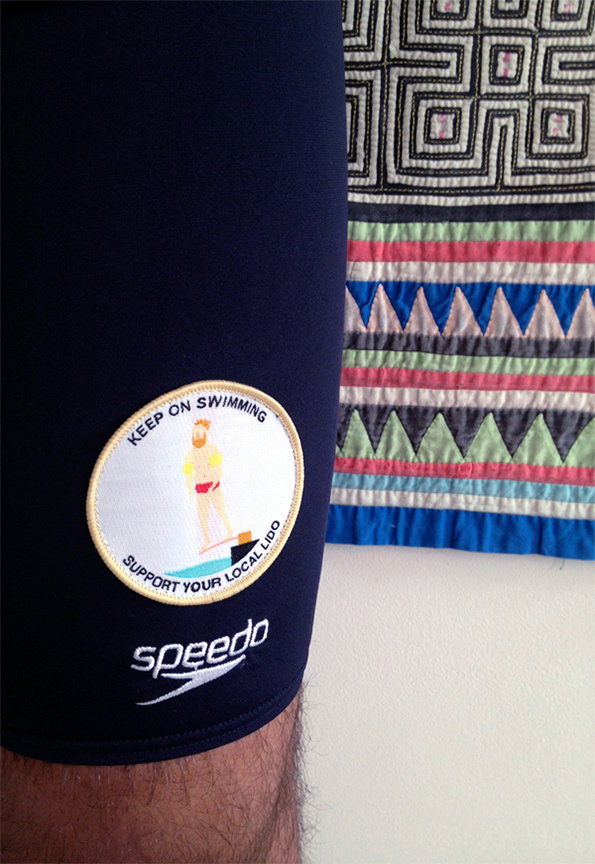 A close-up of a navy blue Speedo swim cap. A circular patch on the side features an illustration of a swimmer and reads “KEEP ON SWIMMING SUPPORT YOUR LOCAL LIDO”.