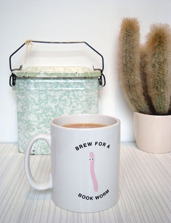 A white mug filled with tea sits on a light surface. It features an illustration of a pink worm and the text “BREW FOR A BOOK WORM”. In the background, a pale green vintage tin and a potted cactus are visible.