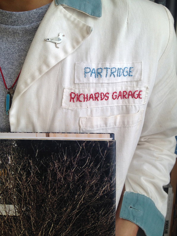 A person wears a white work jacket with blue cuffs and a bird-shaped pin. Two embroidered patches on the chest read “PARTRIDGE” and “RICHARDS GARAGE”.