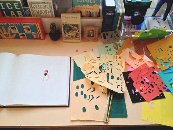 An open sketchbook shows a small cutout of a person on a white page. Beside it, a collection of colourful paper cutouts and a green cutting mat clutter a desk.