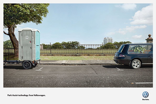 A dark blue Volkswagen car is parked in a bay, positioned perfectly between a portable toilet on a trailer and the end of the parking row. Below the image, text reads “Park Assist technology from Volkswagen.” alongside the Volkswagen logo and the phrase “Das Auto.”.