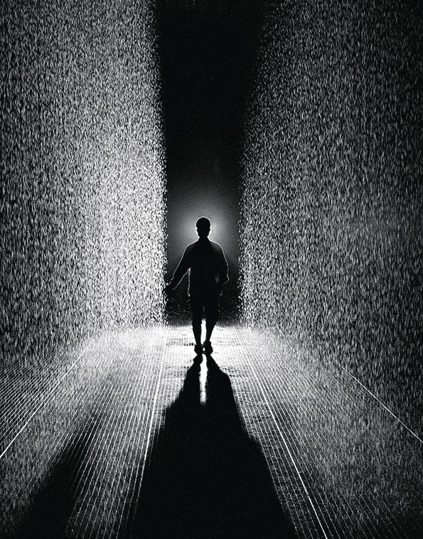 A silhouette of a person stands between two dense walls of falling water. The scene is captured in high-contrast black and white, casting a long shadow across the ribbed floor.