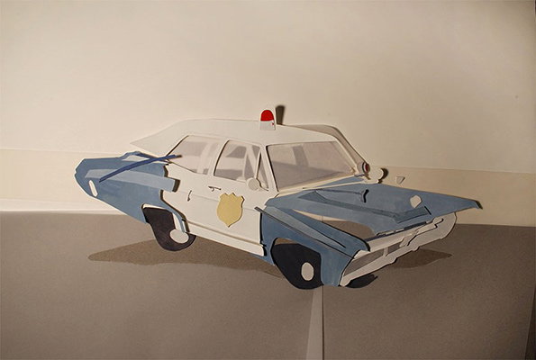A paper model of a police car, constructed from cut and folded card. The vehicle is blue and white with a red siren on the roof and a gold badge on the door.