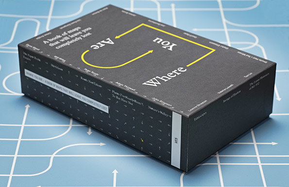 A dark grey book box sits on a blue surface printed with a white grid and arrows. The cover features the words “Where Are You” arranged in a loop, accompanied by the text “A book of maps that will leave you completely lost”.