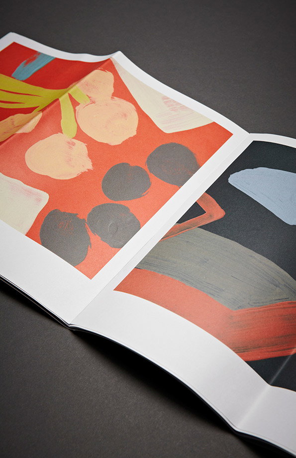 An open book displays two abstract paintings with bold, painterly shapes. The left page features red, yellow and grey forms, while the right page shows a composition of grey, blue and red.