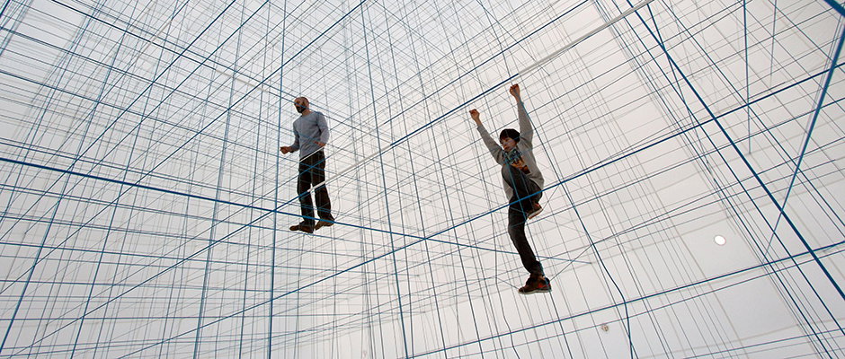 Two people stand within a vast, immersive installation of intersecting blue strings. The lines create a dense, geometric grid that fills the white room from floor to ceiling.