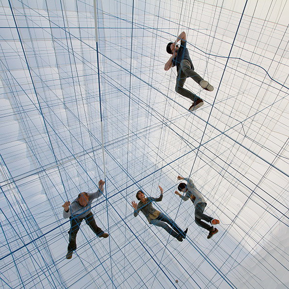 Four people lounge and climb within a vast, three-dimensional grid of blue netting. The structure fills a bright white space, suspending the figures at different heights.