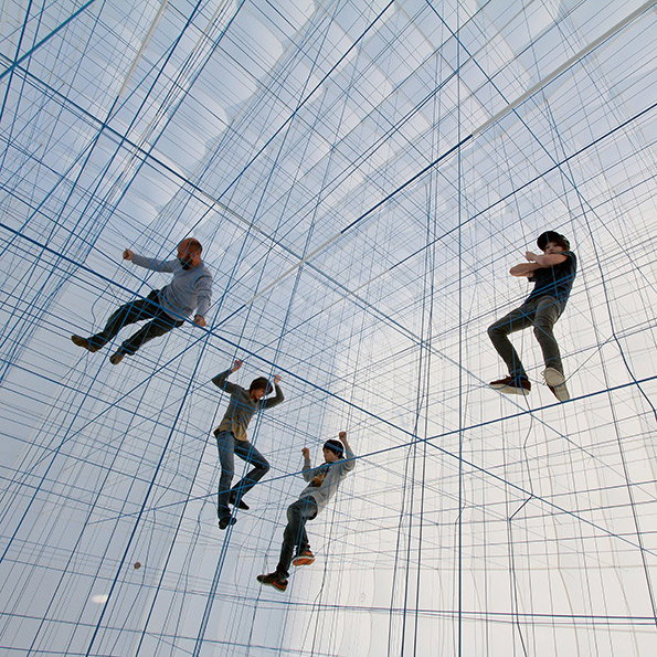 Four people climb and lounge within a vast, three-dimensional grid of blue ropes. The installation fills a bright, airy space, creating a translucent web of intersecting lines.