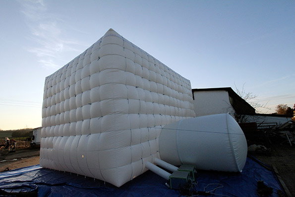 A large, white inflatable cube sits on a blue tarpaulin in an open outdoor space. A smaller, rounded inflatable structure is attached to its side by two white pipes.