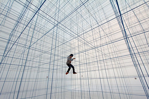 A person leaps through a vast, immersive installation of intersecting blue strings. The lines create a dense, geometric grid that fills the entire white space.
