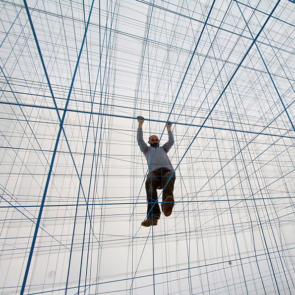 A person hangs suspended within a vast, three-dimensional grid of blue strings. They hold onto a horizontal line with both hands, floating in the centre of the installation.