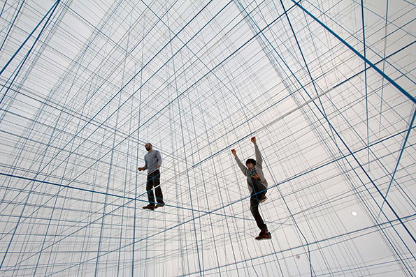 Two people stand within a vast, immersive installation of intersecting blue ropes. The ropes create a dense, three-dimensional grid that fills the white gallery space.