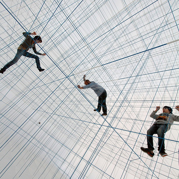 Three people climb and balance across a vast, intersecting grid of blue ropes. The ropes stretch across a bright, white space to create a complex 3D web.