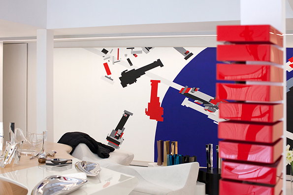 A bright, white interior features a large wall graphic of pixelated red, black and grey shapes against a blue circle. In the foreground, a stack of glossy red rectangular blocks stands beside a white curved sofa and table.