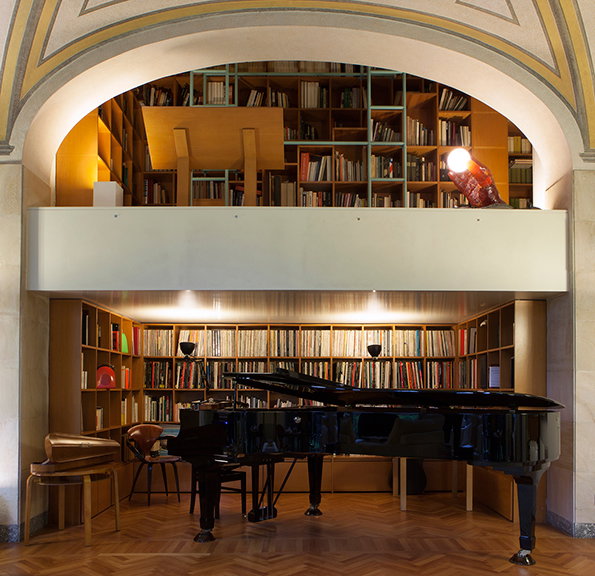 A black grand piano sits in a room with herringbone wood floors. Behind it, floor-to-ceiling bookshelves are packed with books, with a second level of shelving visible through an archway above.