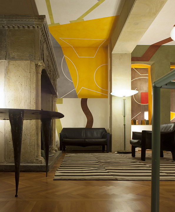 A living room featuring a large stone fireplace and a black leather sofa. The walls and ceiling are decorated with bold, geometric shapes in yellow, red and brown.