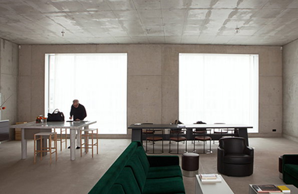 A person stands at a white table in a minimalist concrete room with two large, bright windows. A dark dining table and chairs sit nearby, with a deep green velvet sofa in the foreground.