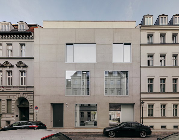 A modern, three-storey concrete building stands between two traditional white European townhouses. The concrete facade features large, rectangular windows that reflect the surrounding architecture.
