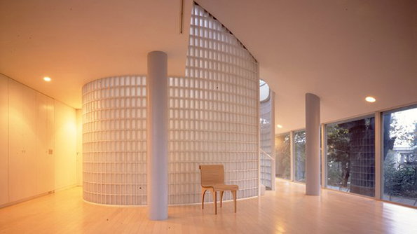 A minimalist interior featuring a curved wall made of translucent glass blocks. A single wooden chair sits on a light timber floor between the wall and a grey support pillar.