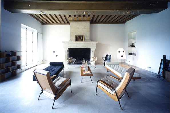 A spacious, minimalist living room with white walls and a dark beamed ceiling. Mid-century modern chairs and a low black sofa surround a small wooden coffee table in front of a large stone fireplace.