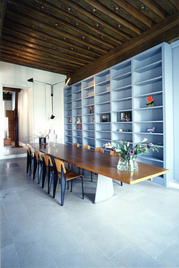A long wooden dining table with black and tan chairs sits in a room with a carved wooden ceiling. Behind the table, a large, light blue built-in shelving unit spans the wall, holding a few scattered objects.