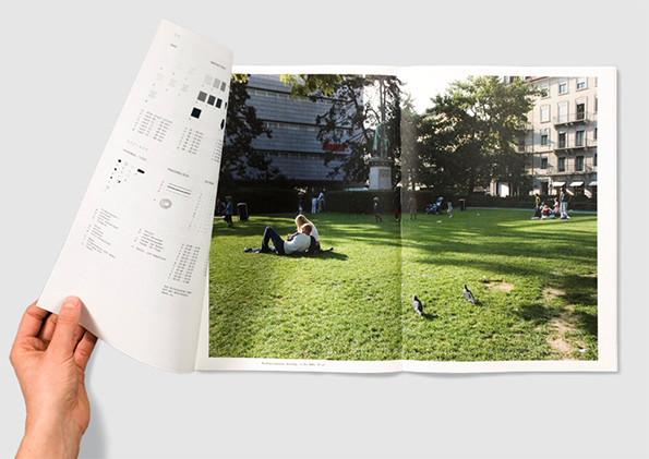 A hand turns the page of an open book to reveal a full-bleed photograph of a sunny park. Two people lounge on a green lawn, with city buildings and leafy trees in the background.