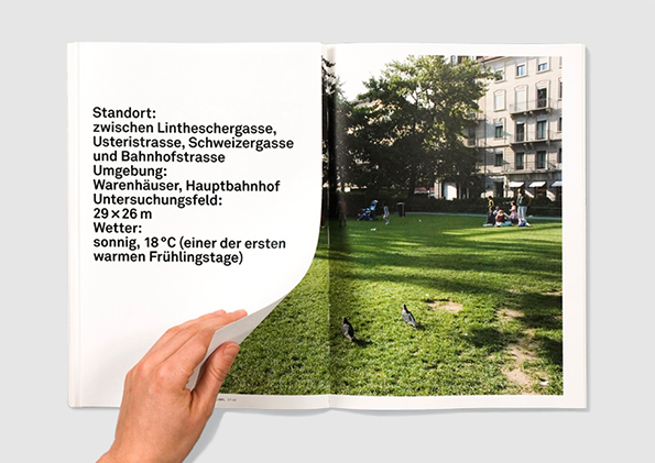 An open book shows a page of German text opposite a photograph of a sunny city park. A hand peels back the bottom corner of the left page to reveal the grass and pigeons in the image beneath.