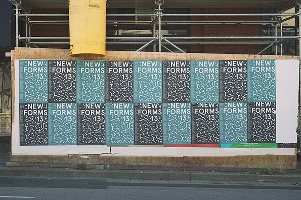 A grid of posters is pasted onto a wooden construction hoarding. Each poster reads “NEW FORMS 13” over a pattern of small, white, pill-shaped marks on a dark teal background.
