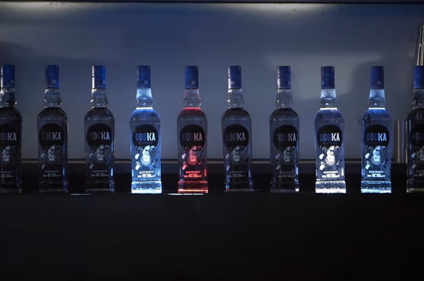 A row of Oddka vodka bottles stands on a dark surface against a dim blue background. Several bottles are lit from below, glowing in shades of blue and red.